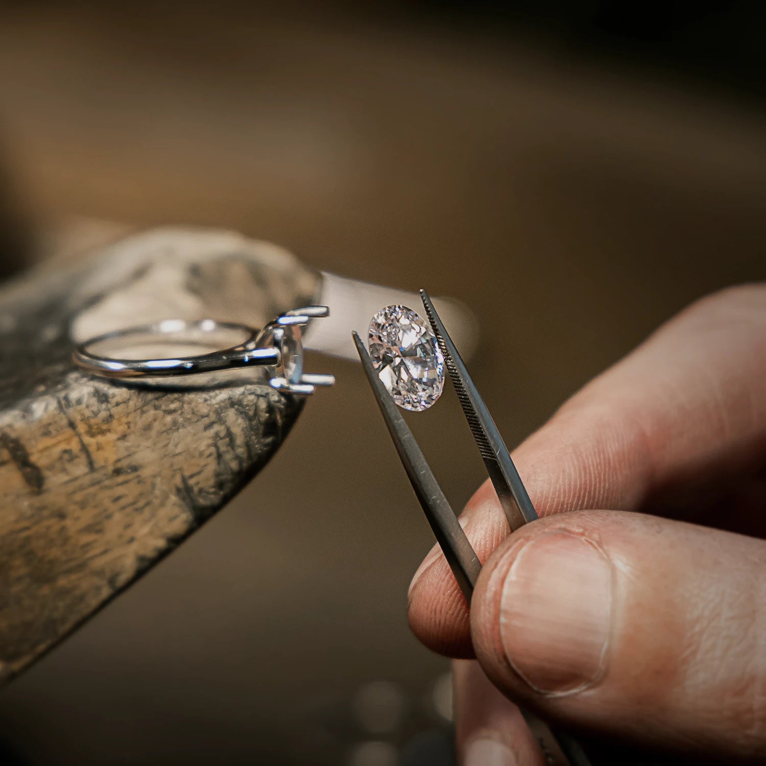 Jeweller holding a loose oval diamond with tweezers next to a ring setting on a workbench