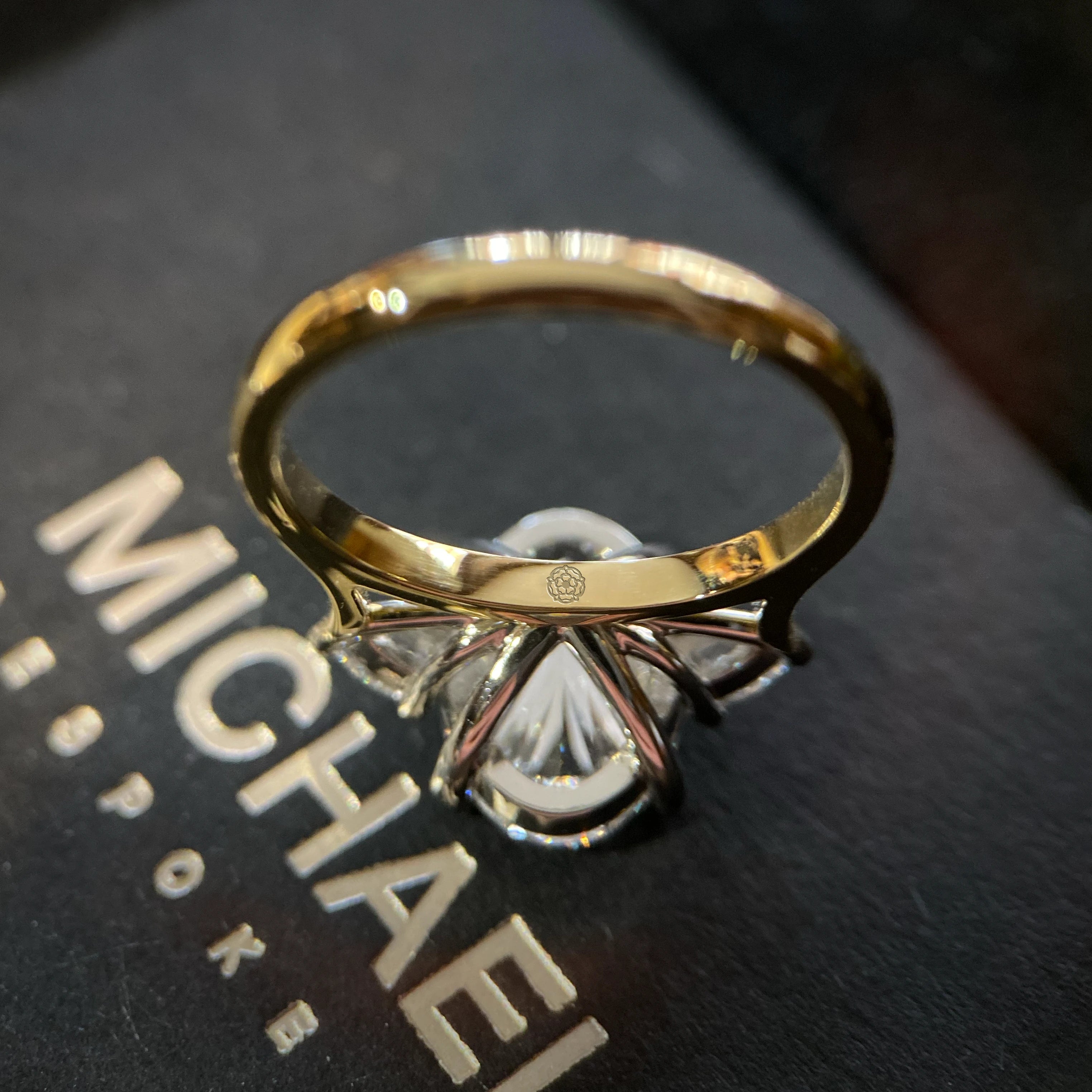 Gold designer engagement ring york with a diamond on a black surface with 'MICHAEL ISAAC' branding.